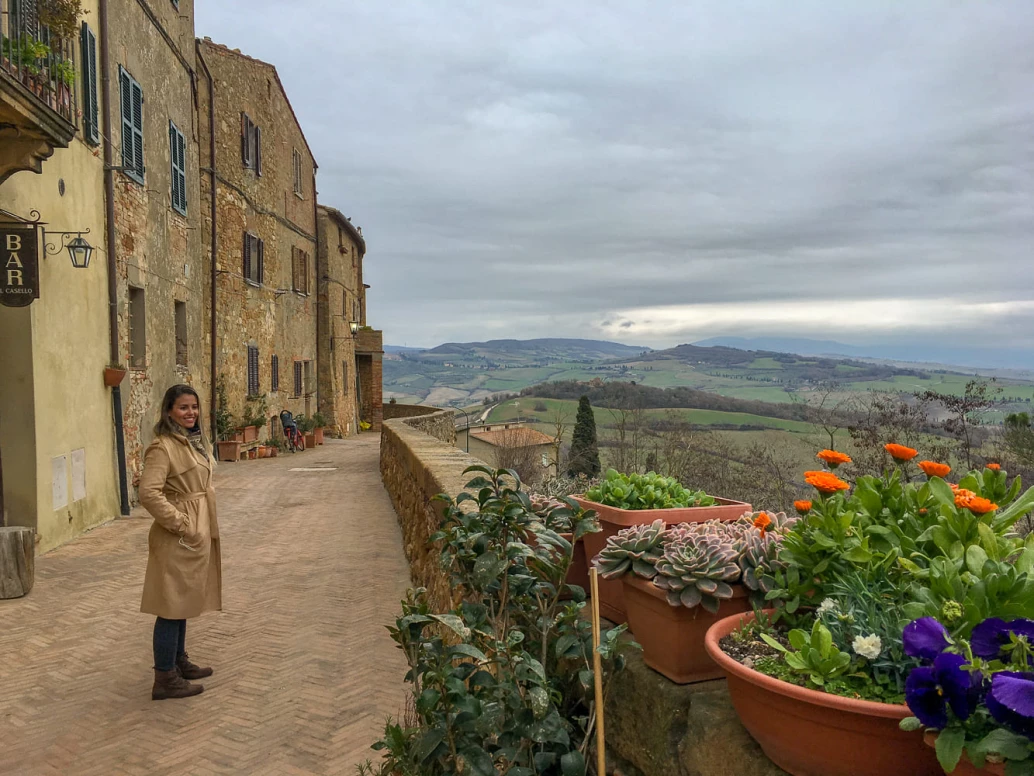 Vista de um burgo na Toscana, Itália. A construção medieval contrasta com a natureza típica e céu densamente carregado. Uma mulher sorri enquanto aprecia a paisagem.
