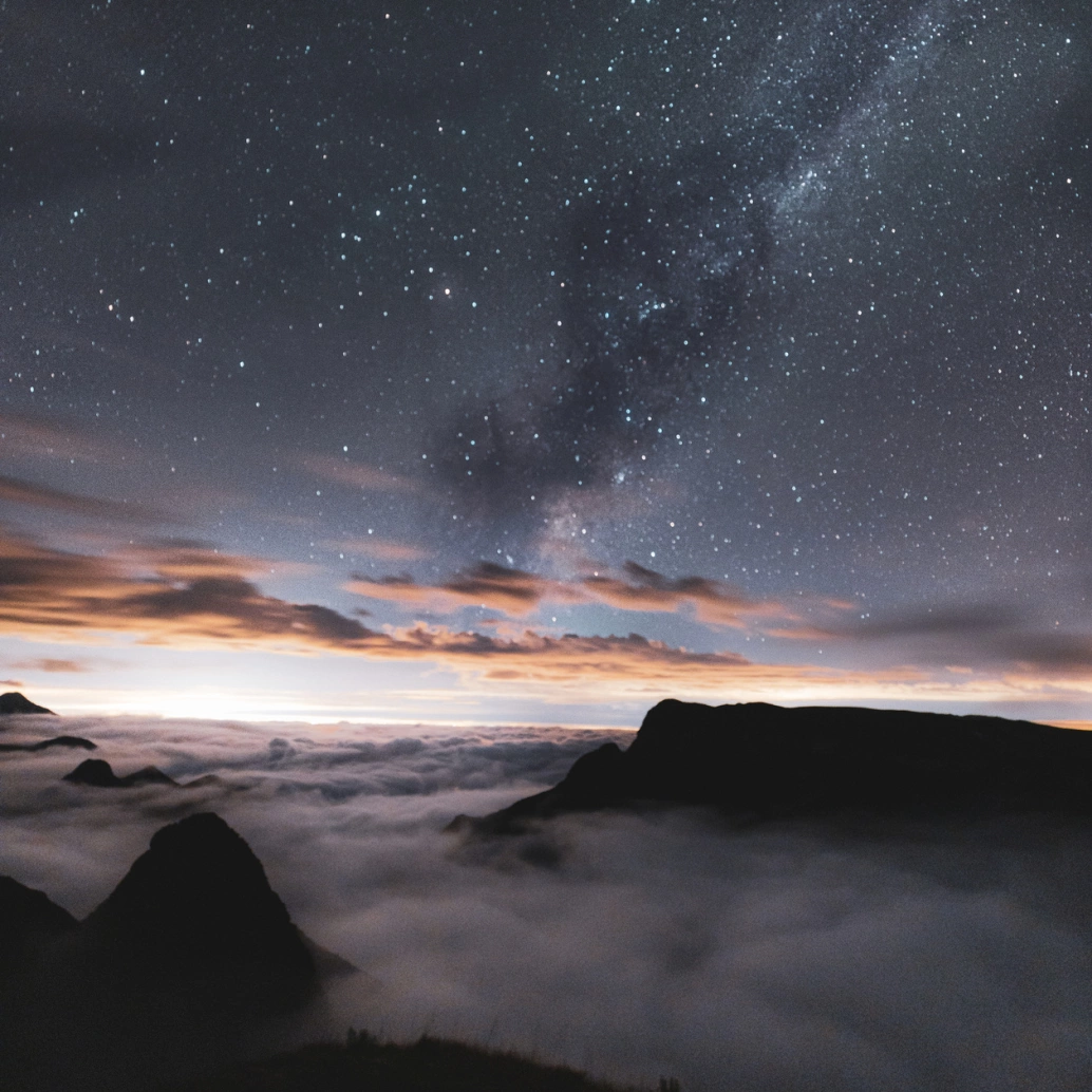 Pontas de penhascos acima de uma camada de neblina. Céu noturno estrelado, com algumas nuvens, através de fotografia de longa exposição