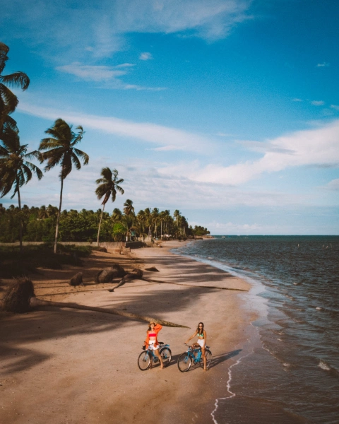 Foto da Praia de Tatuamunha, São Miguel dos Milagres, destacando duas mulheres de bicicleta acenando para a câmera.