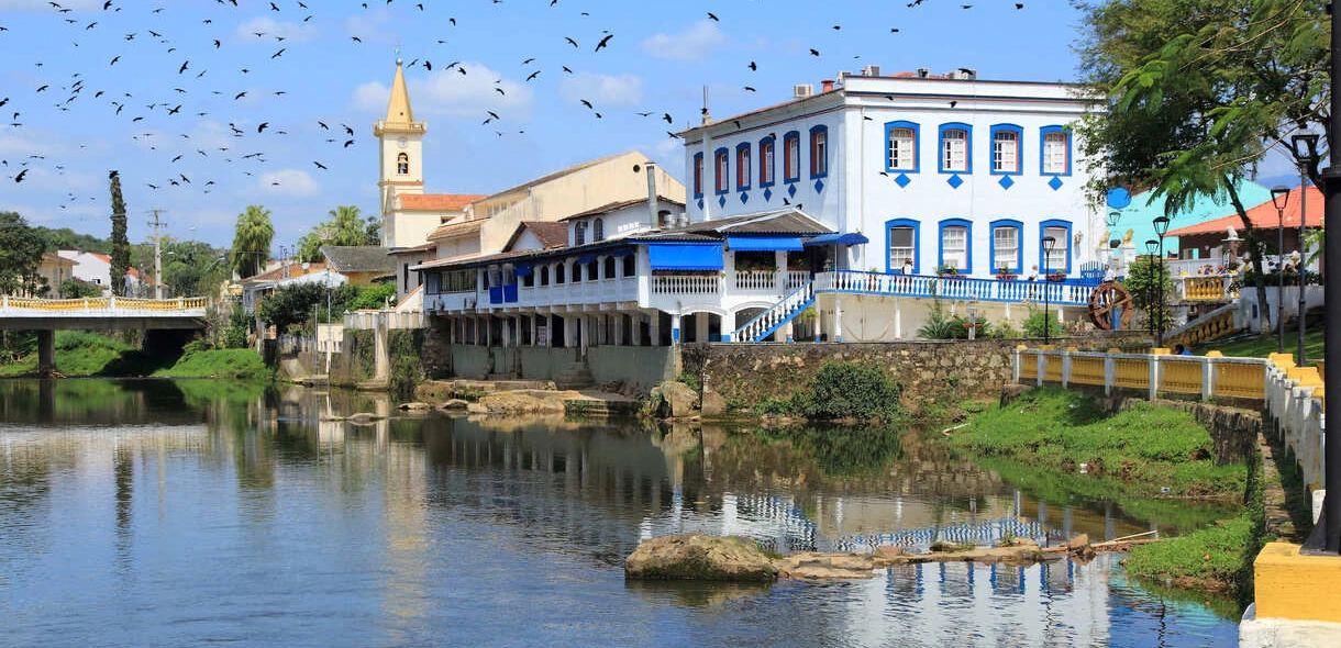 Dia ensolarado com revoada de pássaros sobre um rio que atravessa a cidade. Construções coloniais nas margens do rio em Morretes