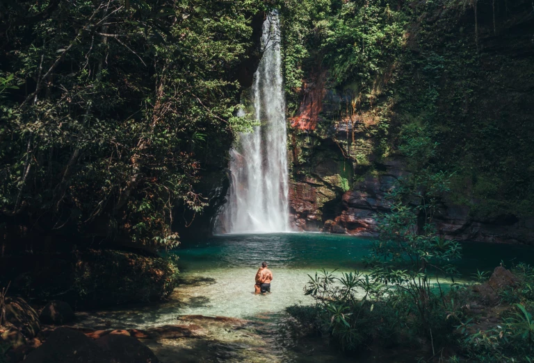 Clareira na mata em Barra das Garças, MT, revela uma linda cachoeira com piscina natural verde-esmeralda cercada de densa vegetação recebendo uma bracha de luz natural em dia claro. Ao centro da imagem, um jovem casal se abraça