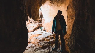 Homem de jaqueta e calça comprida, em pé dentro de uma caverna com equipamentos de segurança e uma lanterna presa à cabeça