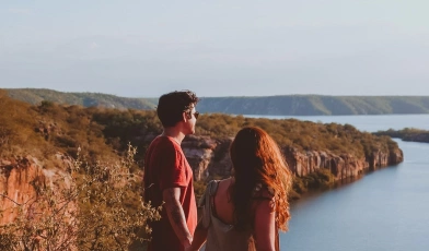 Um homem e uma mulher de mãos dadas contemplam linda paisagem de enorme rio cercado por montanhas em dia ensolarado