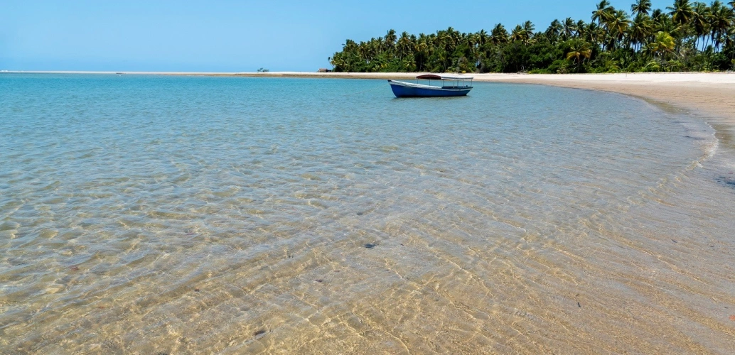 Paisagem de águas rasas e cristalinas de uma praia deserta onde está estacionado um barco e ao fundo pés de coqueiros às margens da areia branca