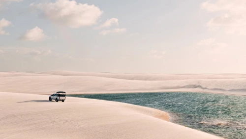 Cenário de um dia ensolarado com um carro passando por dunas que cercam um lago