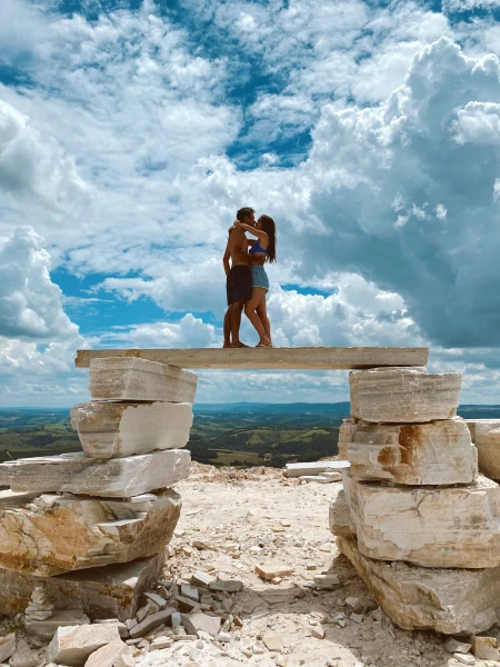 Casal de homem de mulher se beijando em cima de uma plataforma de pedras naturais na cidade de São Thomé das Letras. O local fica em um pico alto da cidade, que dá vista para uma paisagem montanhosa e céu azul com nuvens