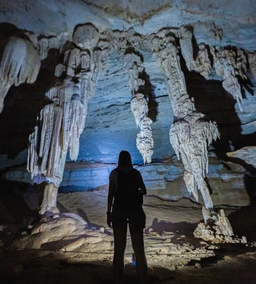Uma mulher de costas ilumina as grutas de uma caverna escura revelando belas formações rochosas