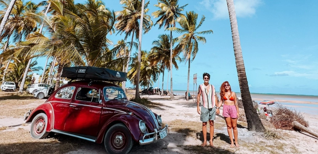 Um homem e uma mulher de mãos dadas ao lado de um Fusca vermelho estacionado nas areis de uma praia deserta com vários coqueiros ao fundo