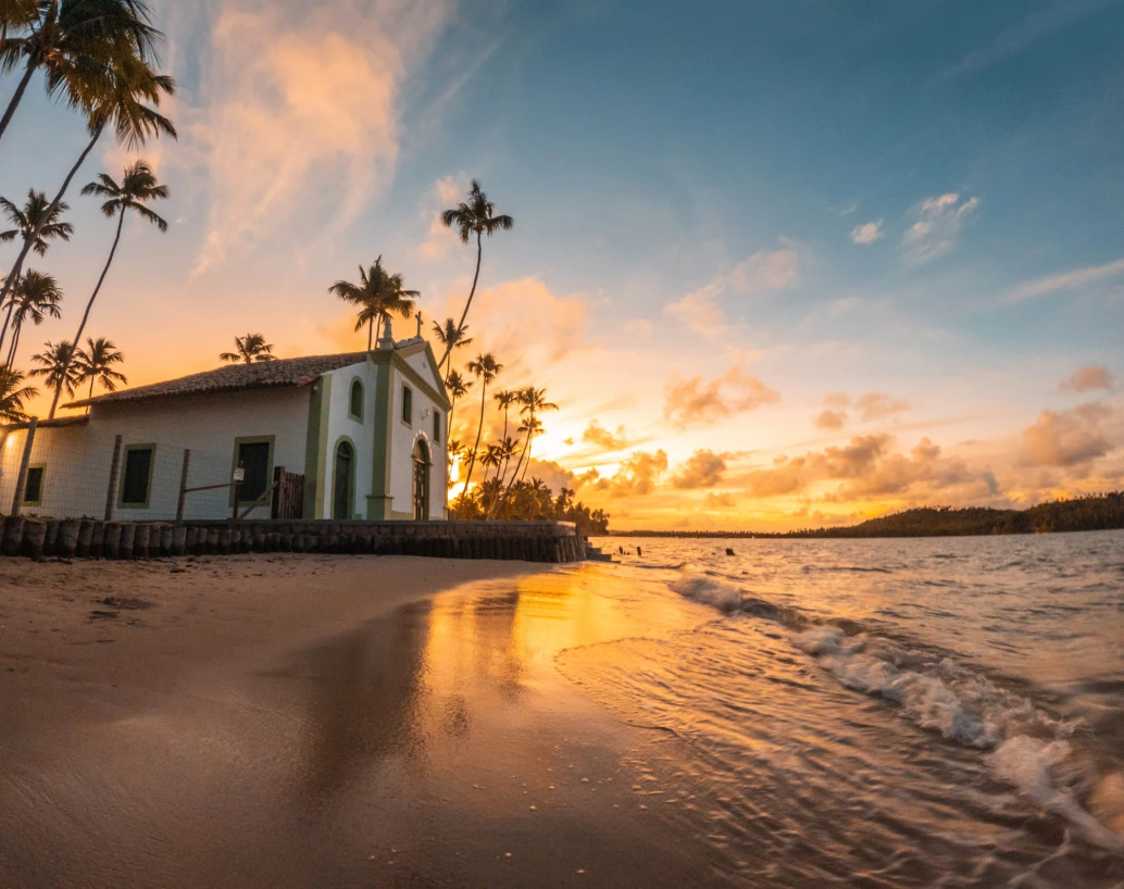 Pôr do sol na Praia de Carneiros. O sol se põe atrás de uma capela, refletindo sua tonalidade laranja na areia e marcando a silhueta dos coqueiros.