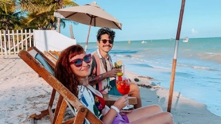 Uma mulher e um homem tomam um drink sentados na beira de uma praia deserta em dia ensolarado