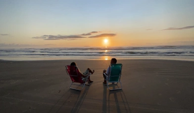Homem e mulher sentados em cadeira de praia assistem nascer do sol