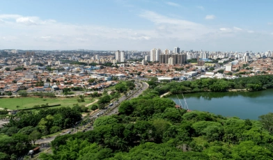 Vista aérea de cidade com bastante natureza. Uma lagoa se destaca no plano frontal e, ao fundo, há casas e prédios