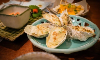 Em uma mesa de madeira escura, um prato de cerâmica azul claro com ostras gratinadas. Em segundo plano, outros recipientes com outros tipos de comida.