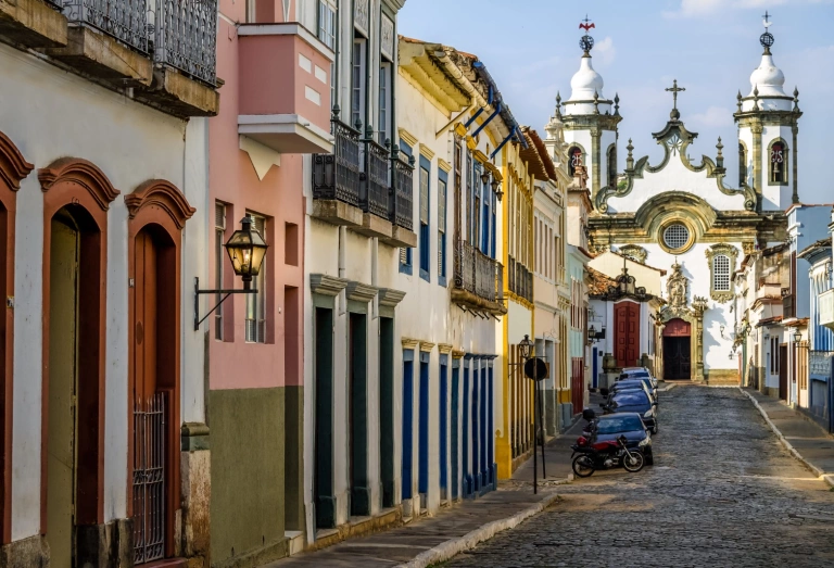 Vista de igreja do período colonial numa cidade histórica em Minas Gerais, São João del Rei. A frente se destaca uma rua com casas antigas.