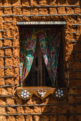 Janela de uma casa simples construída com tijolos de barro e vigas de madeira. Há uma cortina florida e, no topo, uma placa escrito “Lavras Novas MG”