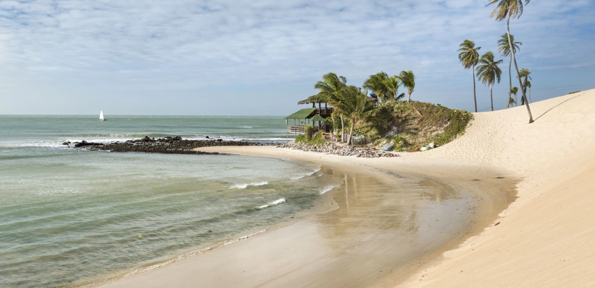 Vista de praia paradisíaca e deserta, mar esverdeado e dunas de areia clara se destacam.