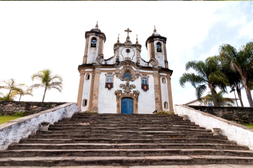 Fachada de uma igreja de arquitetura colonial sob degraus de uma grande escada cercada por árvores