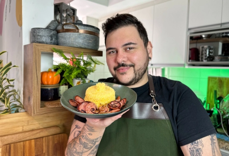Um homem branco, de cabelos pretos, cavanhaque e tatuagens pelos braços em um cozinha, segurando um prato de cerâmica contendo cuscuz nordestino com linguiça calabresa e cebolas.