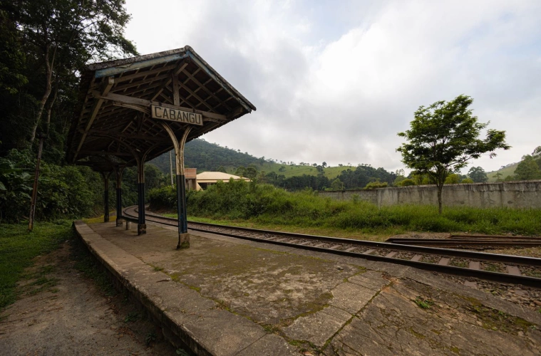 Pequena estação de trem com estrutura simples e placa escrito “Cabangu”. O trilho faz uma curva e, ao fundo, há muita vegetação