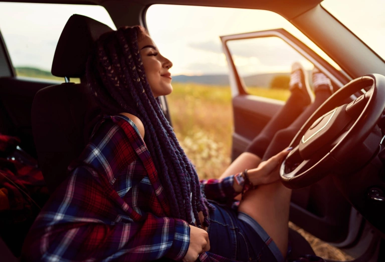 Mulher sentada no banco da frente de um carro com a porta aberta. Ela sorri, está de olhos fechados, segura o volante com a mão esquerda e está com os pés na janela do carro.
