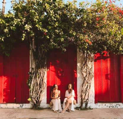 Duas mulheres posam para foto sentadas em calçada. Ao fundo portas vermelhas e plantas de muro.