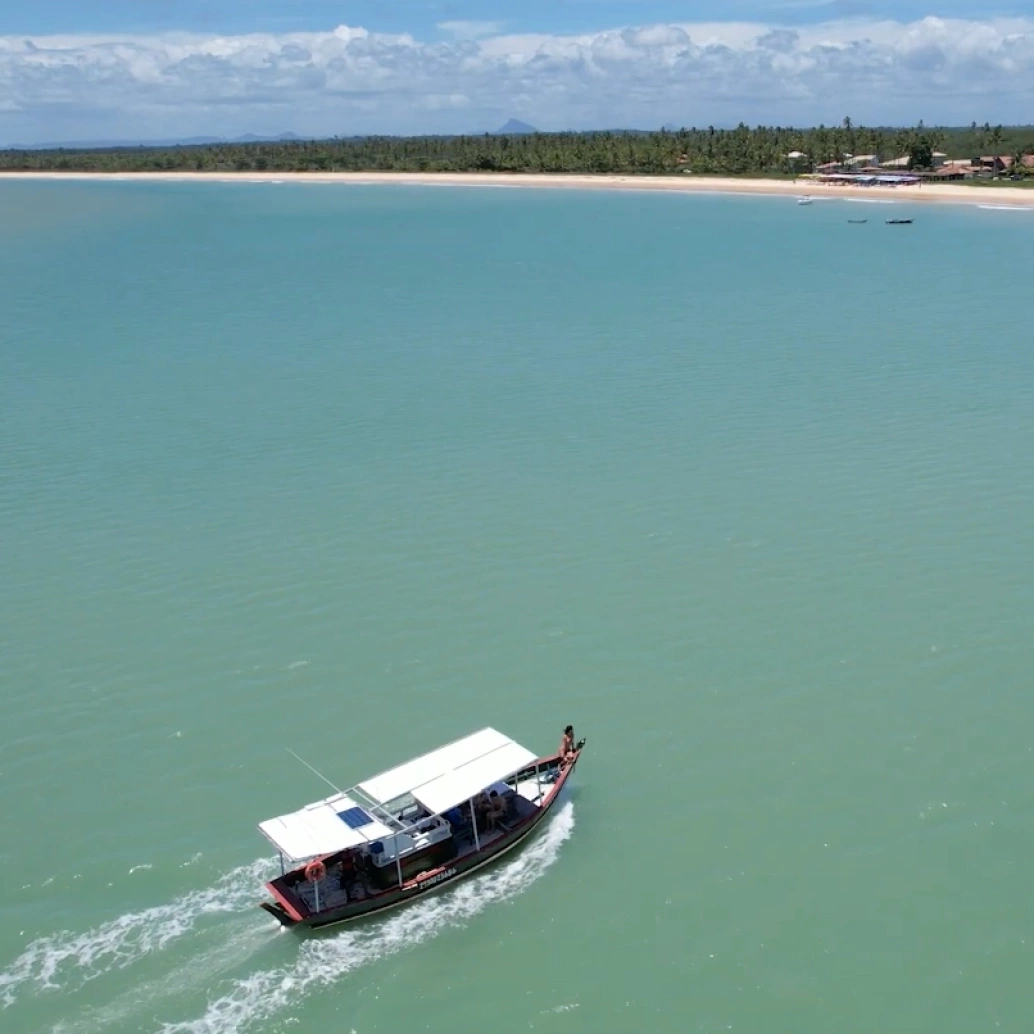 Vista aérea de uma praia de águas de tons verde e azul cristalinos com um barco navegando por ela em dia ensolarado