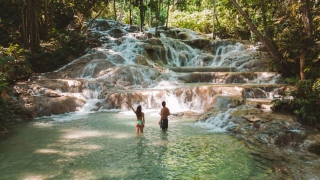 Mulher e homem de costas com meios corpo no poço da cachoeira enquanto apreciam a queda d’água que escorre em meio às pedras