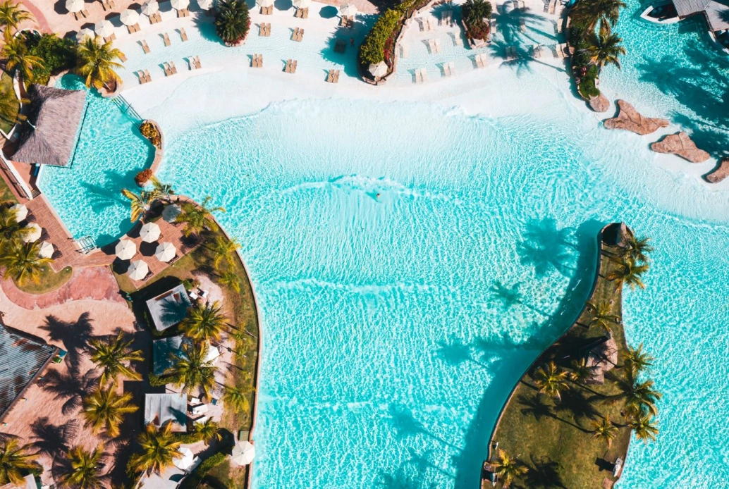 Fotografia aérea da piscina artificial com pequenas ondas, cercada de coqueiros, e alguns quiosques. Espreguiçadeiras enfileiradas ao longo da orla.