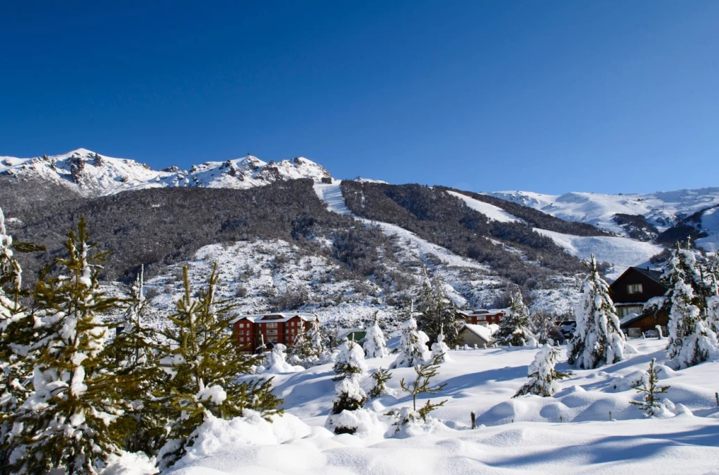 Vista panorâmica de uma paisagem coberta de neve nas montanhas de Bariloche, com pinheiros e um hotel ao fundo.