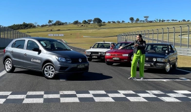 Quatro gerações do Gol Volkswagen juntos em um autódromo em um dia ensolarado. Com eles, uma mulher de jaqueta e blusa preta, calça verde cítrico.