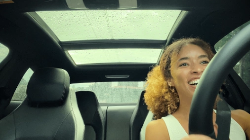 Garota sorrindo com mãos no volante dentro de carro