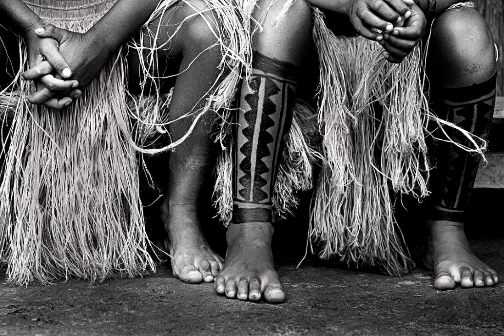 Foto em preto e branco, destacando as pernas e mãos de duas pessoas indígenas, com suas vestes e tatuagens típicas.