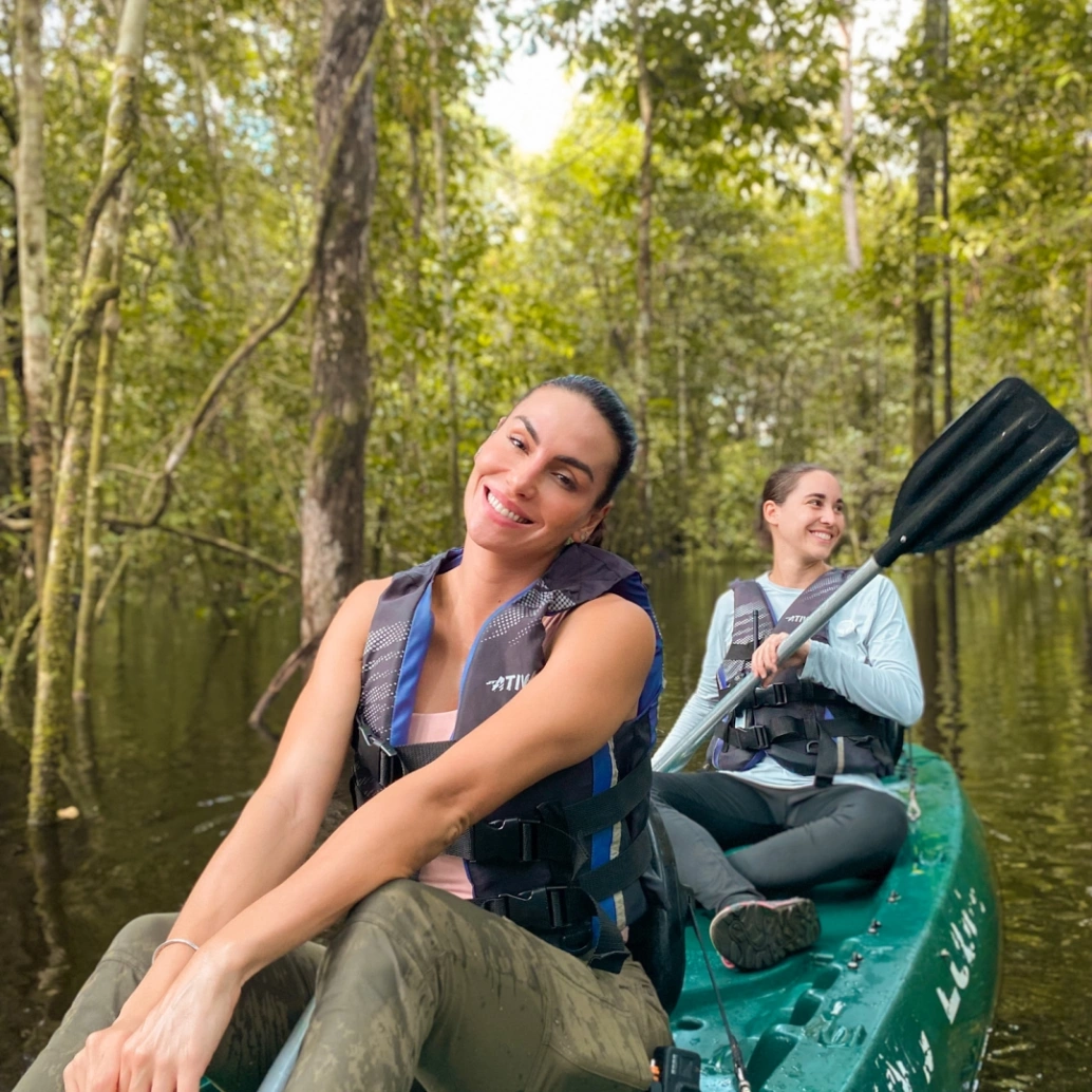 Mel Fronckowiak e a guia turística, Jéssica, sorridentes em uma canoa em meio à Floresta Alagada.
