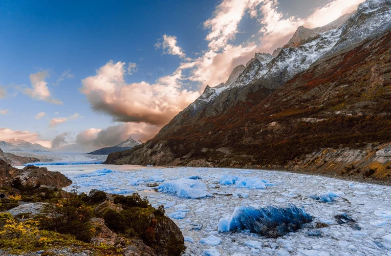 Lago com placas de gelo em sua superfície, em meio à paisagem da Patagônia chilena. Há relevos com vegetação nas margens, e montanhas nevadas ao fundo