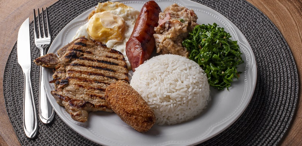 Prato branco com arroz, couve, feijão, linguiça, ovo frito e bife suíno. Ao lado, uma faca e um garfo prateados. Ambos sobre uma mesa de madeira.