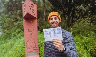 Homem jovem com jaqueta e touca sorrindo para a câmera e segurando um passaporte. Ele está ao lado de um marco da Estrada Real, com vegetação ao fundo