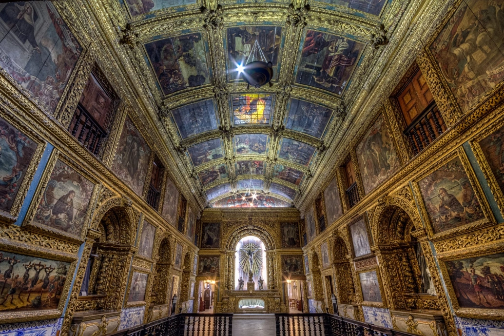 Interior de uma igreja ornamentada com ouro e pinturas sacras nas paredes e no teto. O altar aparece ao fundo