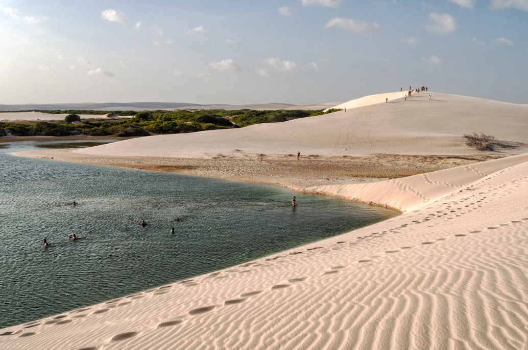 Em um dia ensolarado, dunas de areia cercam um lago com alguns banhistas