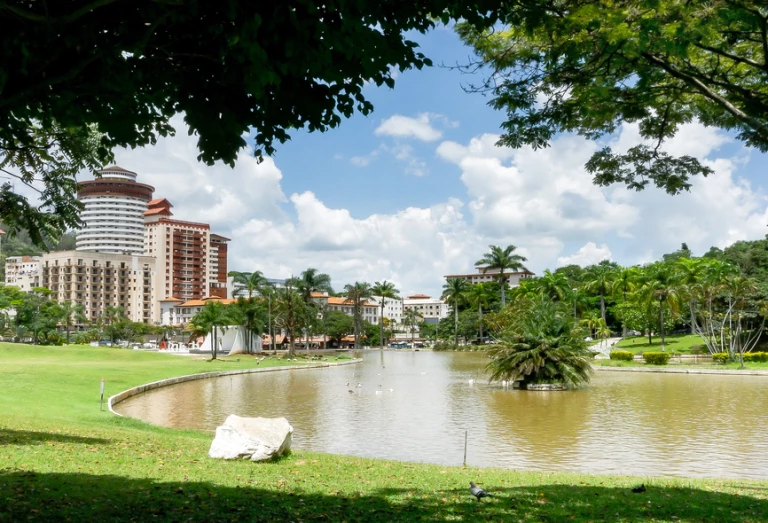Praça arborizada com um lago no meio, na cidade de Águas de Lindóia, SP. As margens, e em meio à grama bem aparada, a vegetação típica da Mata Atlântica se destaca. Ao fundo, algumas construções de classe média-alta adornam a paisagem num dia claro