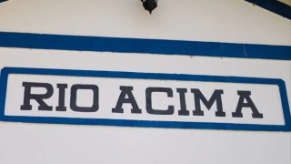 Foco em uma placa escrito “Rio Acima” em uma construção colonial em tons de branco e azul