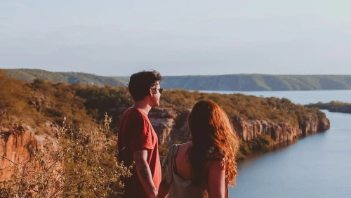 Um homem e uma mulher de mãos dadas contemplam linda paisagem de enorme rio cercado por montanhas em dia ensolarado