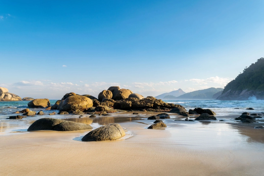 Conjunto de pedras esféricas sedimentadas em praia, no encontro das águas azuis do mar e areia em dia claro