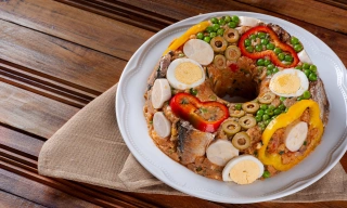 Em uma mesa de madeira clara, um prato branco com cuscuz paulista, decorado com ovos, pimentões, ervilhas, sardinhas e azeitonas.