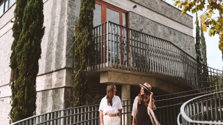 duas mulheres posam para foto na frente de um restaurante em Bento Gonçalves - RS. Há uma escadaria branca, com o restaurante ao fundo com parede de pedras.