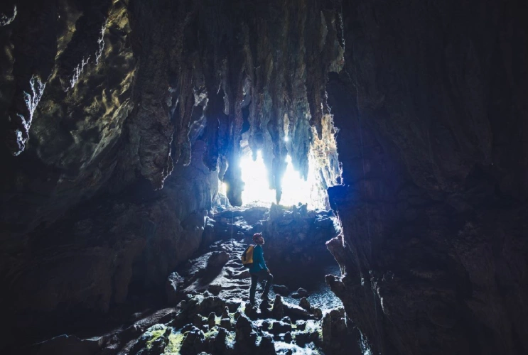 Homem em pé em meio às estruturas de uma caverna escura, com luz entrando por uma fenda e iluminando as estalactites no teto