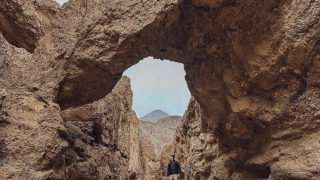 Homem em cima de uma pequena pedra em meio a uma cânion entre duas montanhas rochosas. Acima dele, uma ponte natural formada pelas pedras