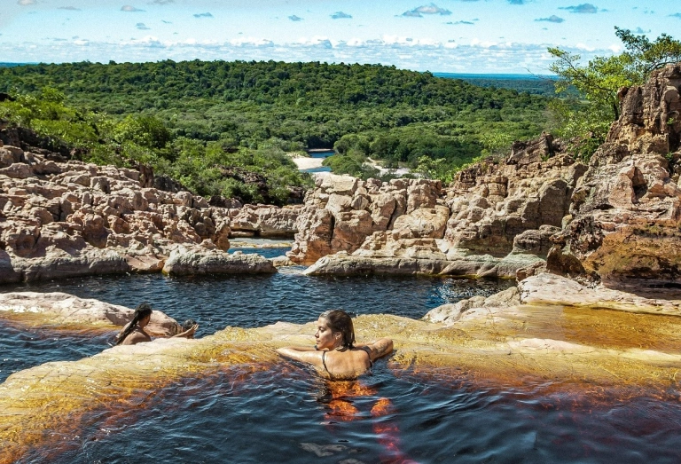 Duas pessoas nadam em piscina natural de uma cachoeira cercada por rochas e vasta vegetação preservada em dia claro