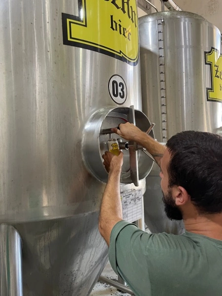 Rapaz de cabelos escuros e curtos tirando cerveja de dentro de um barril de alumínio em uma cervejaria.