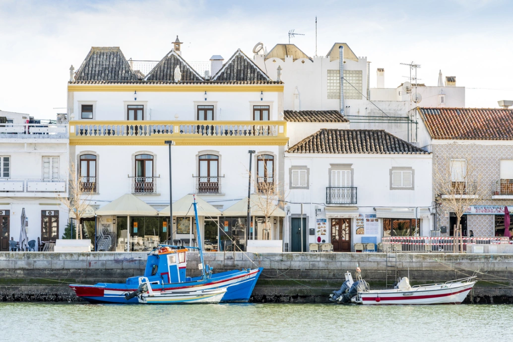 Dois barcos ancorados na margem do Rio de Gilão. Ao fundo, arquitetura tradicional de Tavira no Algarve, Portugal.