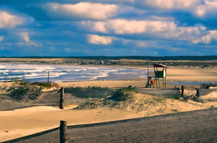 Vista lateral de praia em dia nublado. Uma extensa faixa de areia por toda a imagem e, em primeiro plano, uma cadeira típica de salva-vidas